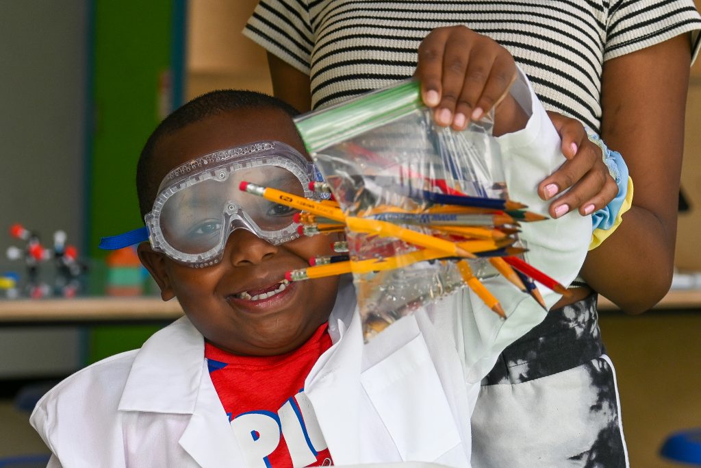 Child wearing goggles and a lab coat, holding up a bag filled with water and punctured with pencils for a science experiment.